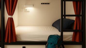 Camera con letto a castello, un letto singolo, un tavolino con una lampada, una presa di corrente e uno zaino per terra.