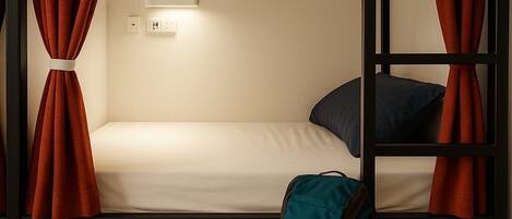 A bunk bed room with a single bed, a small table with a light, a power outlet, and a backpack on the floor.