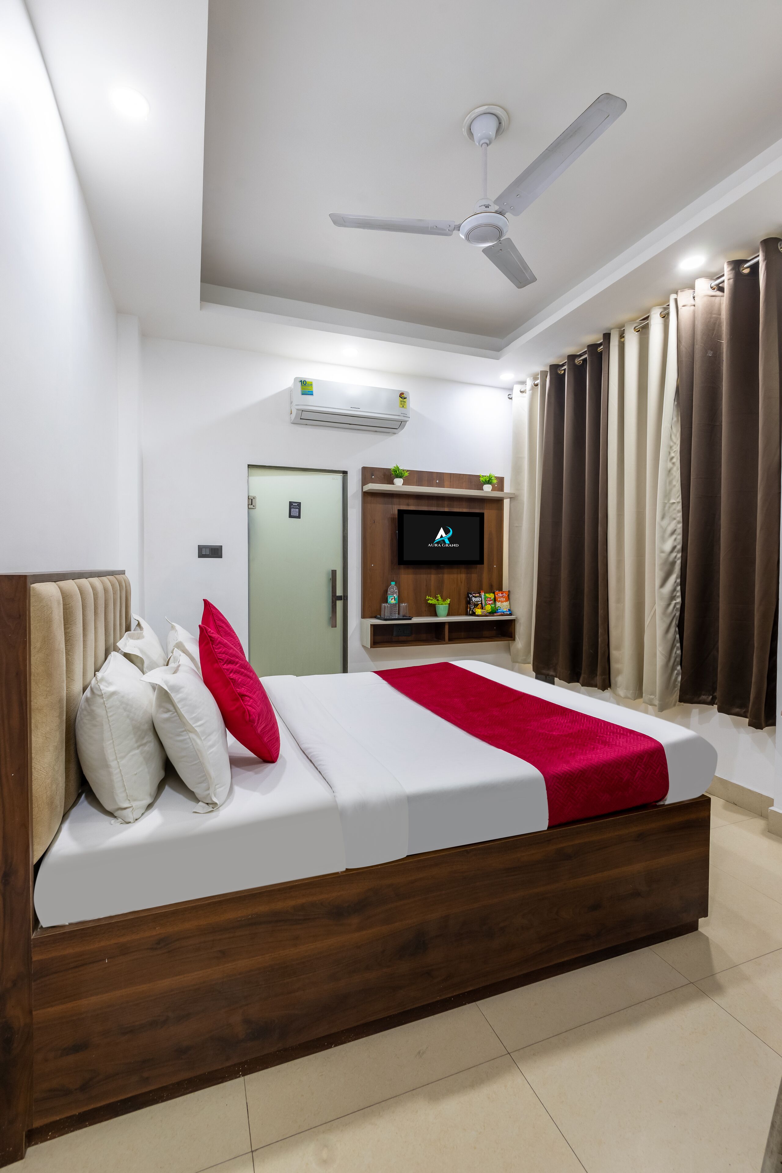 Una habitación de hotel moderna con una cama de madera, ropa de cama blanca, un plaid rojo, un ventilador de techo, un televisor y un estante pequeño con artículos de aseo.