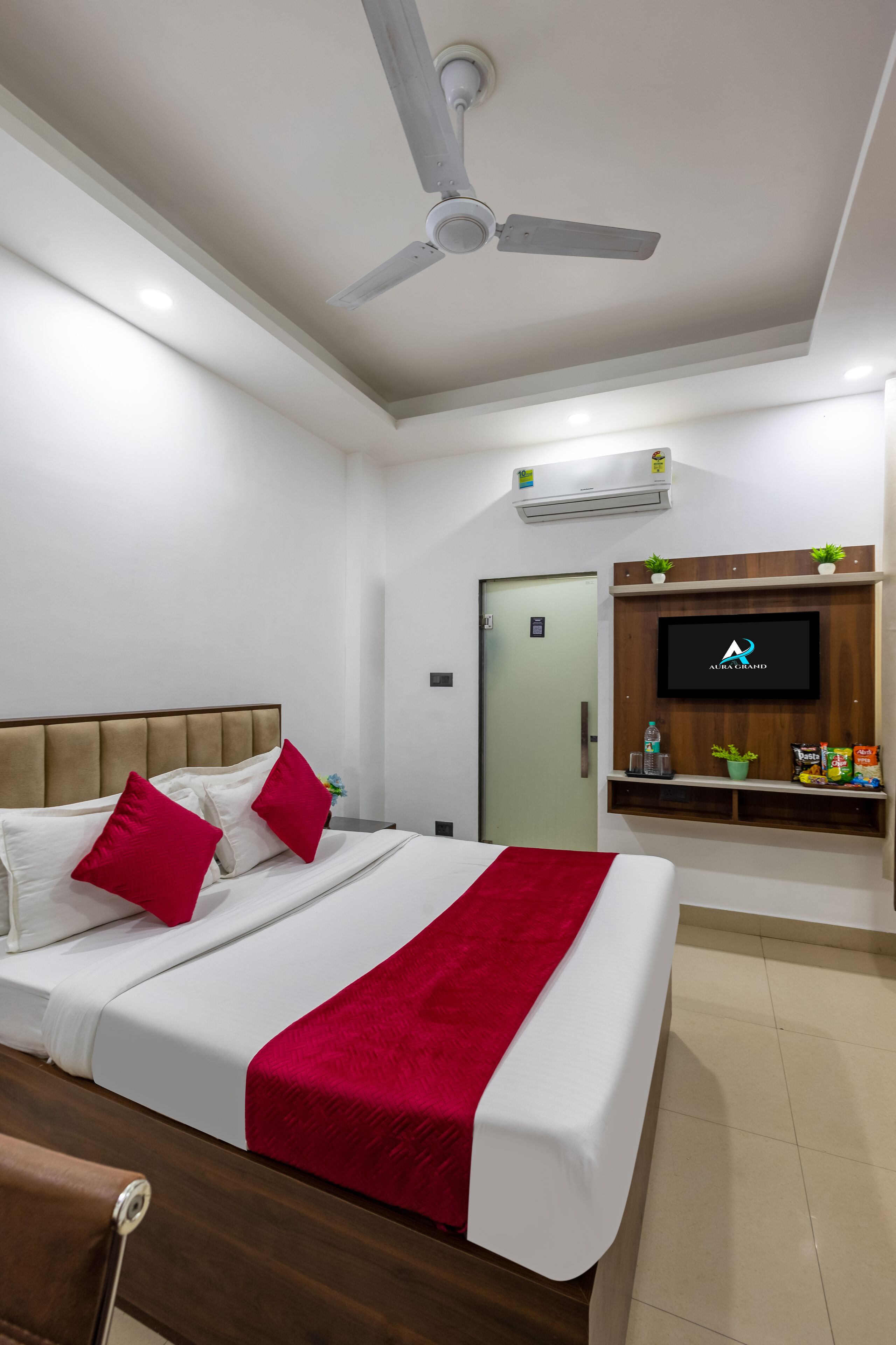 A hotel room with a bed, a ceiling fan, a TV mounted on the wall, and a small shelf with snacks.
