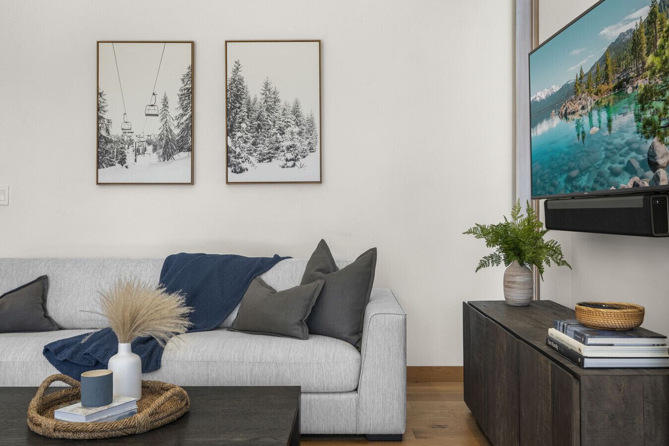 Een woonkamer met een grijze bank, een salontafel met een vaas en boeken, een televisie op een houten dressoir, en twee ingelijste foto’s aan de muur.