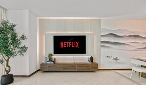 A modern living room with a flat-screen TV mounted on a wall, a wooden console table with books, a potted plant, and a large wall mural. - WYZ RENT|Creekside Haven 2BR Marsa Plaza DFC (Dubai)