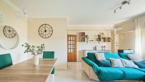 A modern living room with a wooden dining table, teal sofa, and two wall clocks.