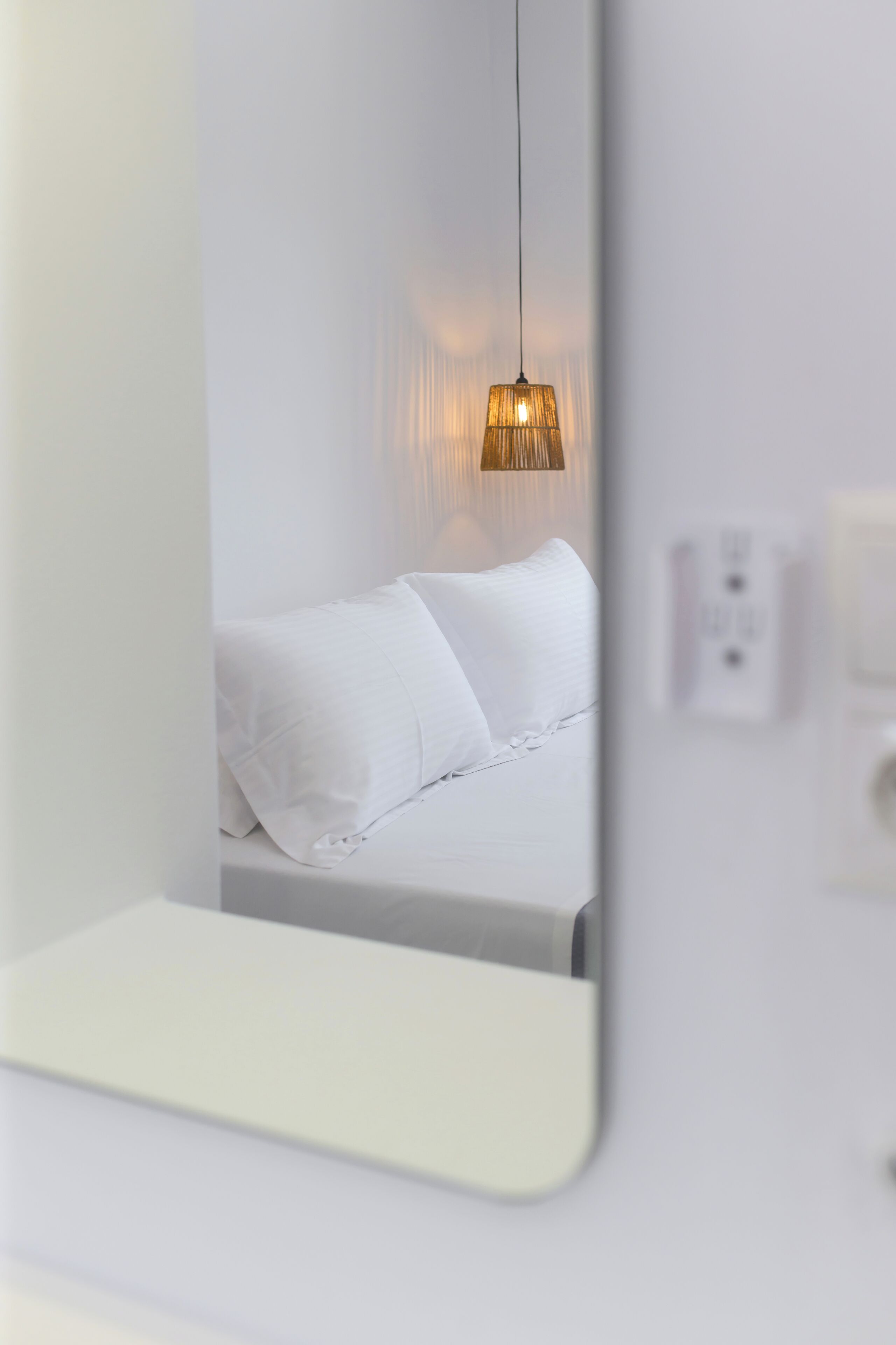 Un espejo refleja una cama con ropa de cama blanca y una lámpara colgante.