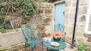 Un bâtiment en pierre avec une porte bleue, un petit jardin avec une table et des chaises, et une plante en pot.