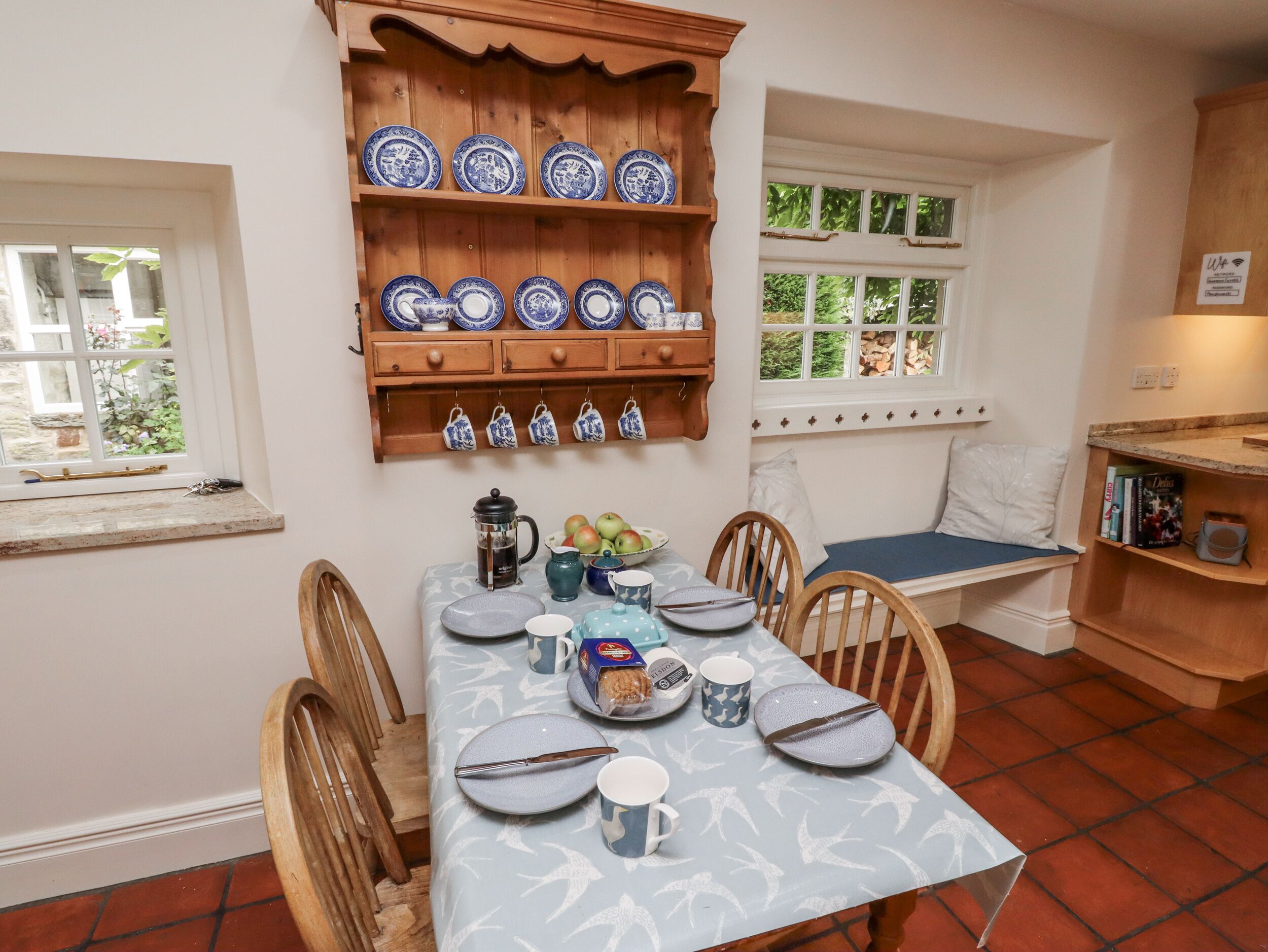 A dining area with a table set for two, a wooden shelf above with blue and white plates, and a window with a view of greenery.