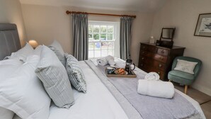 Un lit bien fait, avec des oreillers, un plateau avec une cafetière et des viennoiseries, un fauteuil vert et une commode.
