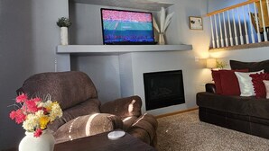 Un salon avec un téléviseur posé sur une étagère, un ventilateur de plafond, une cheminée et un escalier.