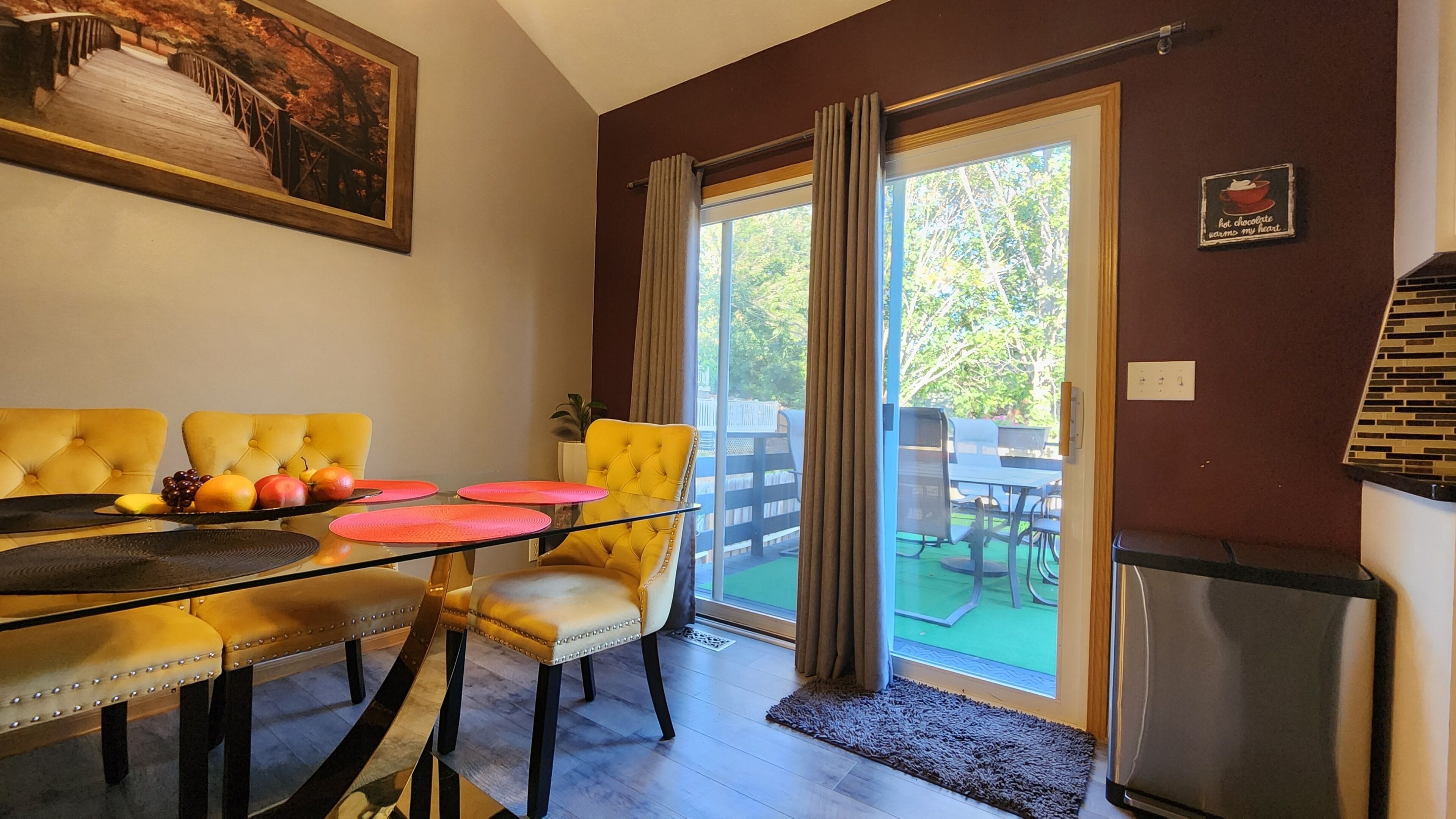 Un coin repas avec une table en verre, des chaises recouvertes d’un tissu jaune et un plateau de fruits.