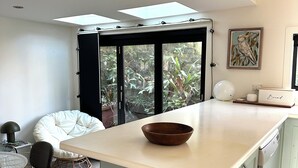 A modern kitchen with a central island, a dining area with a round table and chairs, and a view of greenery through a sliding door. - Large architect designed home directly overlooking the Royal National Park (Bundeena)