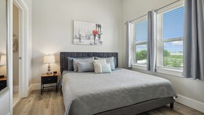 A bedroom with a large bed, a nightstand, a lamp, a window with curtains, and a wall-mounted picture.