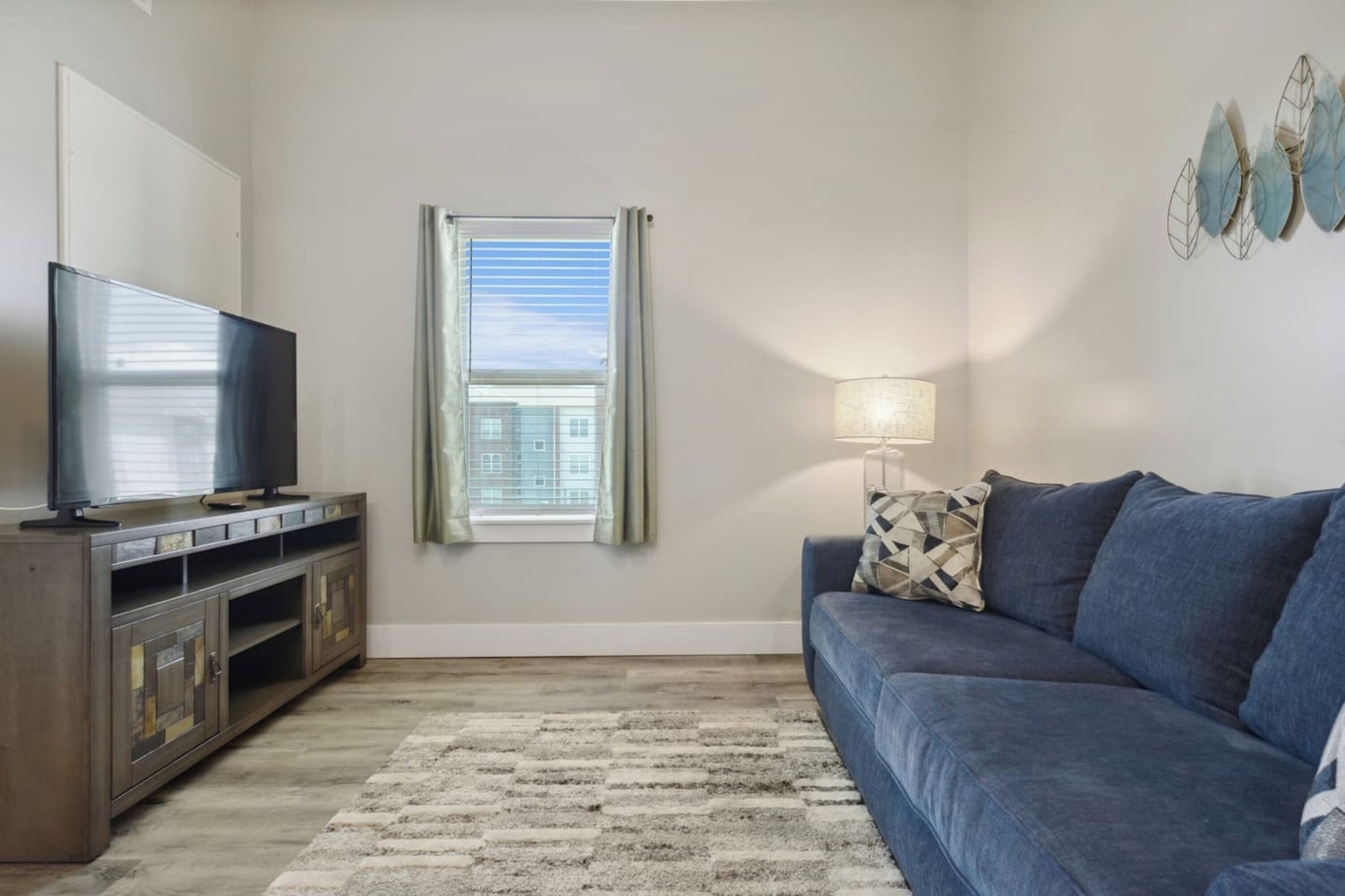 Sala de estar con televisor de pantalla plana sobre un mueble de madera, un sofá azul, alfombra estampada, ventana con cortinas y lámpara fijada a la pared.