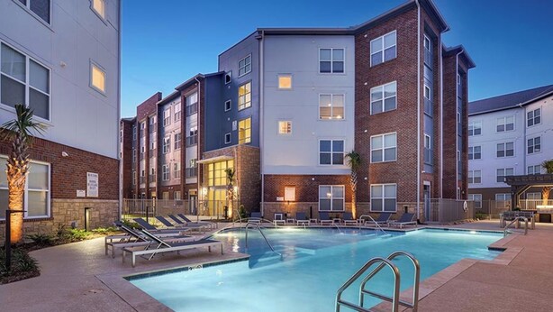 A modern apartment building with a swimming pool, lounge chairs, and a well-lit exterior.