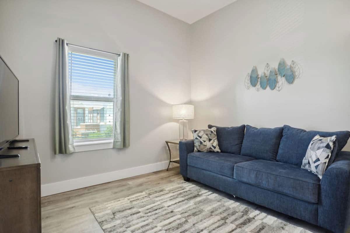 A living room with a blue sofa, a patterned rug, a lamp, and a window with curtains.