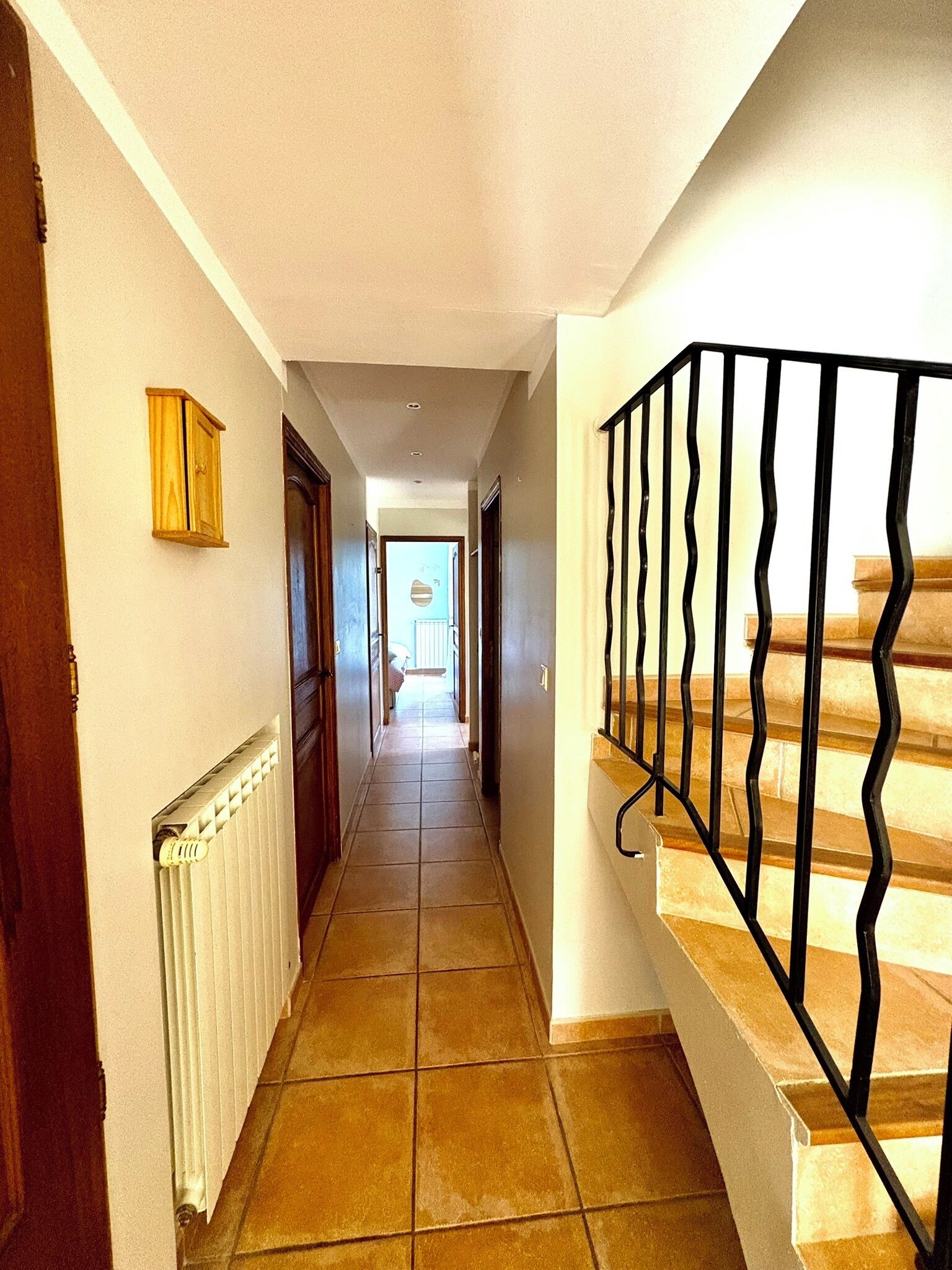 A hallway with a tiled floor, a radiator, a wooden cabinet, and a staircase with a black railing.