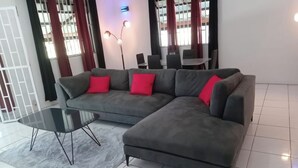 Ein modernes Wohnzimmer mit einem grauen Ecksofa, einem Glastisch und roten Kissen. Eine Stehlampe mit drei Leuchten, ein Esstisch mit Stühlen und ein Blick auf ein Gebäude durch große Fenster mit roten Vorhängen.