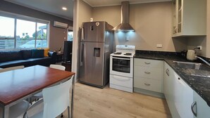 A modern kitchen with a stainless steel refrigerator, oven, and sink. There is a dining area with a wooden table and white chairs. - Cosy Cabin in the Loop (Te Anau)