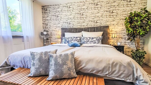 Een slaapkamer met een metselwerk muur, een bed met gekleurde kussens, een ventilator naast het bed, een lamp en een bloempot.