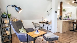 Een moderne woonkamer met een zwarte stoel, een houten salontafel en een zwarte vloerlamp. Er is een keukenhoek met een houten aanrecht en barstoelen.