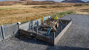 Ein Spielplatz mit Rutsche und Klettergerüsten, umgeben von einem Holzzaun und Kiesgrund, vor einer Hügellandschaft und bewölktem Himmel.