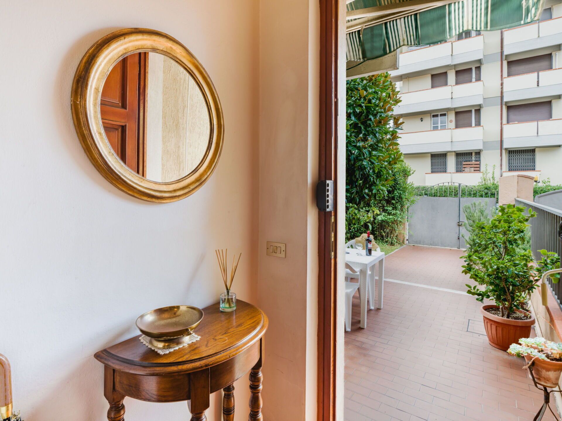 A room with a round mirror, a small wooden table with a bowl and reed diffuser, and a view of a balcony with potted plants and a building in the background.