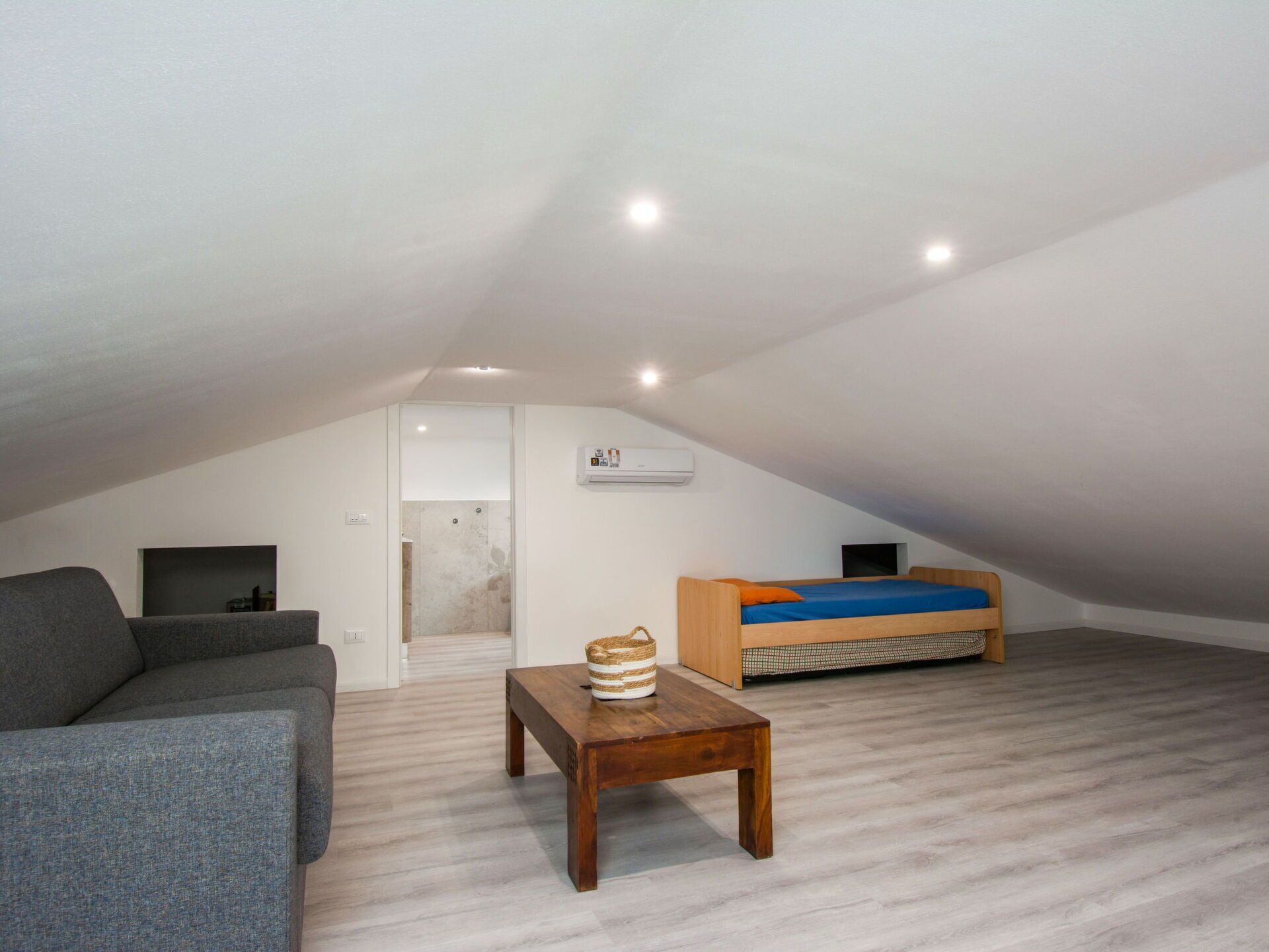 Une petite chambre mansardée moderne, avec un plafond en pente, un lit intégré, un canapé gris, une table en bois et une petite salle de bain avec douche.