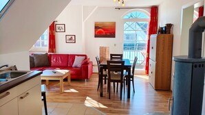 A cozy living area with a red sofa, wooden dining table, and chairs. A kitchen area with a sink and cabinets is visible. A large window with red curtains allows natural light.