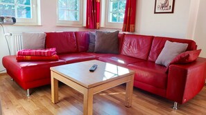 A red leather sofa with a wooden coffee table in a room with wooden flooring, a radiator, and windows with red curtains.