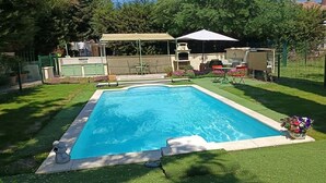Una piscina rectangular con un deck perimetral, una zona de estar cubierta y una parrilla.