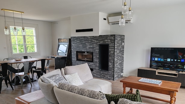 A modern living room with a sectional sofa, a dining table with chairs, a fireplace, a television, and a large window.