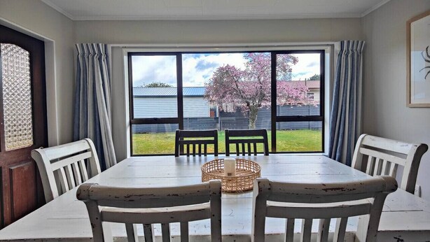 A dining area with a large table and four chairs, a wicker basket on the table, and a view of a body of water and cherry blossoms through large windows. - Luxmore Lookout Te Anau (Te Anau)