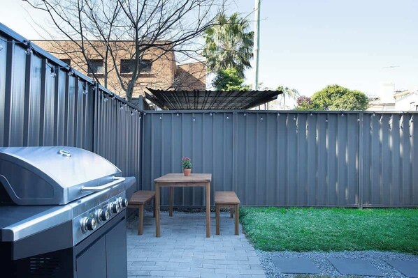 A barbecue grill, a wooden table with two benches, and a fenced backyard.