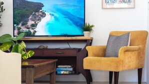 Een woonkamer met een flatscreen televisie op een houten standaard, een gele fauteuil, een salontafel en een boekenkast.