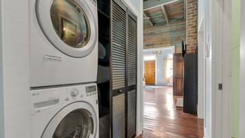 A laundry area with stacked washers and dryers, a wooden floor, and a view into a room with a door and a chandelier.