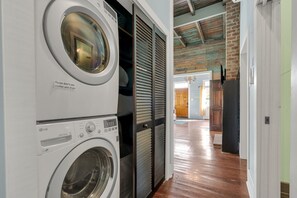 A laundry area with stacked washers and dryers, a wooden floor, and a view into a room with a door and a chandelier.