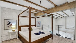 Un dormitorio con cama con dosel, paredes de ladrillo blanco, vigas de madera, una mesita de noche pequeña y un cuadro enmarcado en la pared.