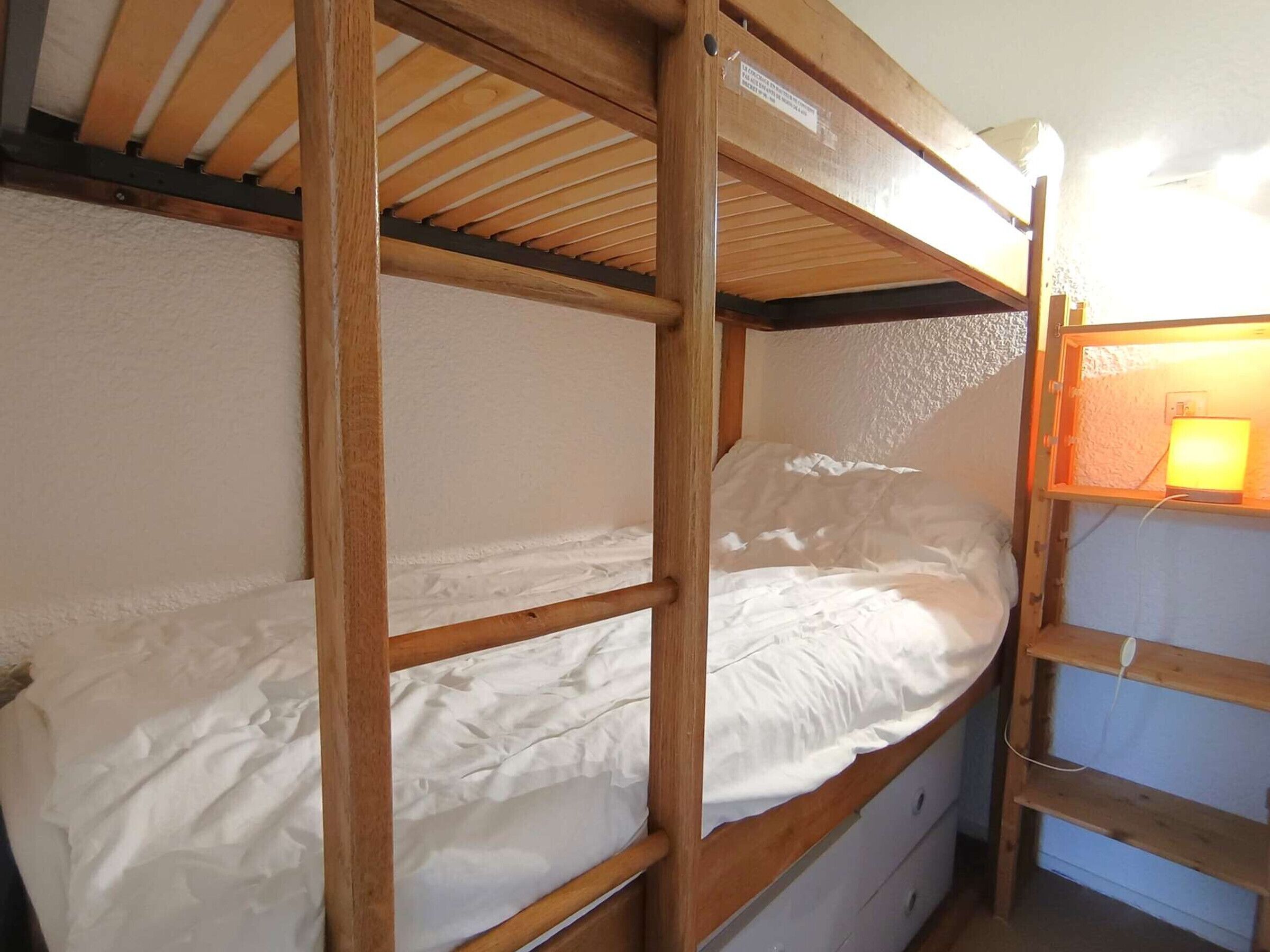 A bunk bed room with a wooden frame, white bedding, and a small bedside table with a lamp.