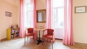 Ein Zimmer mit zwei Holzstühlen, einem kleinen runden Tisch, einem Bücherregal und einem Fenster mit rosa Vorhängen.