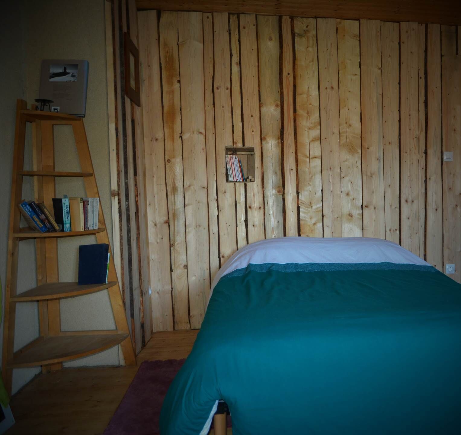 Een slaapkamer met een houten wand, een bed met een blauwgroene sprei en een houten ladderplank.