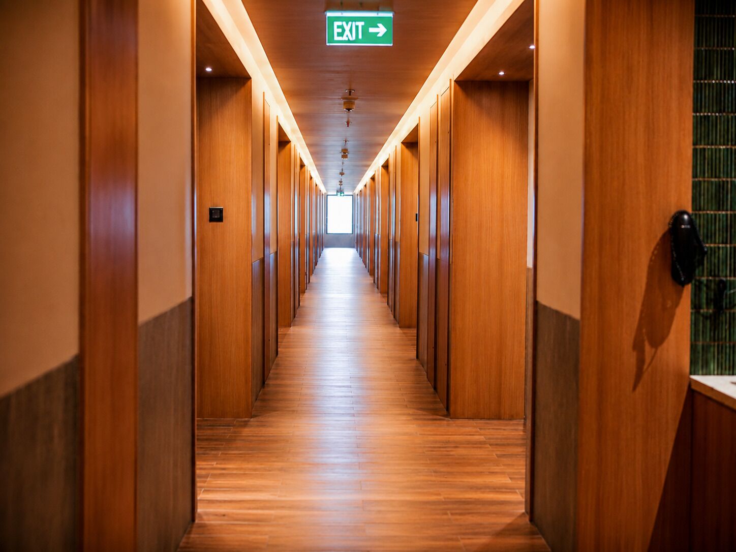 A hotel corridor with wooden walls and a wooden floor, leading to an exit sign.