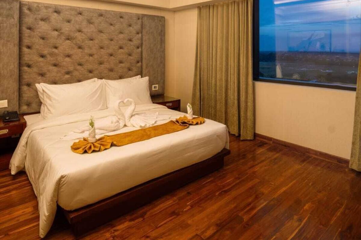 Une chambre d’hôtel avec un grand lit, un plancher en bois, une tête de lit capitonnée, une table de chevet, une fenêtre avec des rideaux et une vue sur l’extérieur.