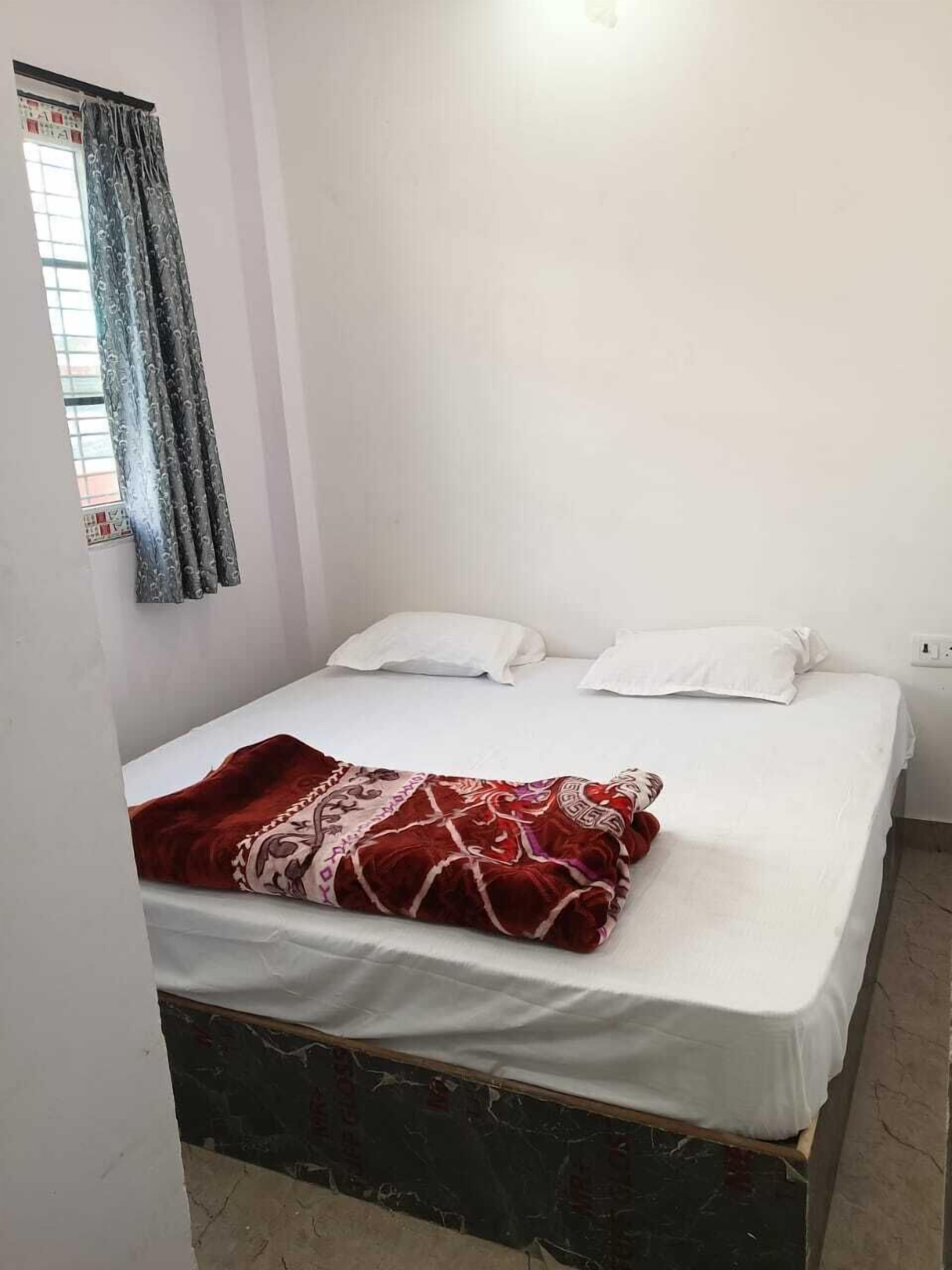 A single bed with white bedding and a red patterned blanket, a window with curtains, and a plain wall.