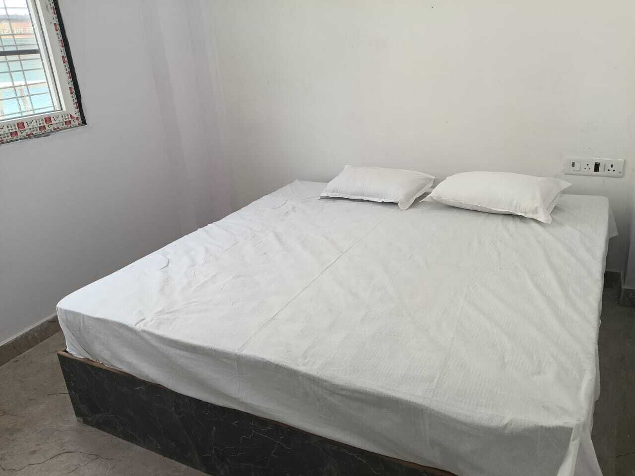 Un lit simple avec un matelas et des oreillers blancs, une tête de lit en marbre, une fenêtre avec des stores et une prise de courant au mur.