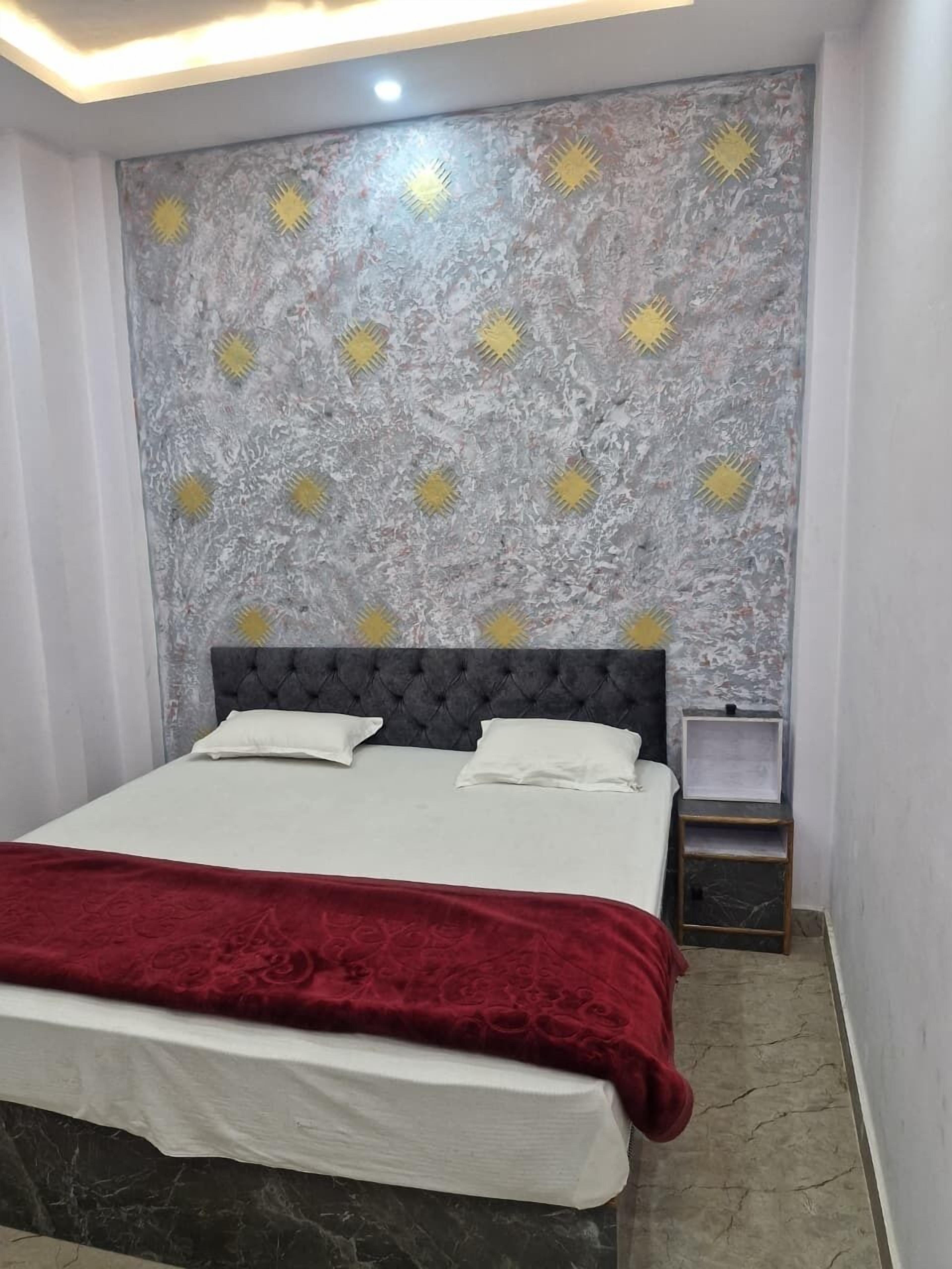 A bedroom with a bed, a red throw blanket, a marble floor, a grey headboard with yellow sun-like patterns, and a small bedside table.