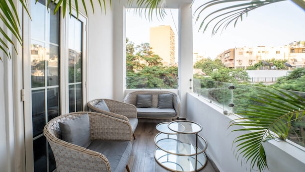 A balcony with wicker furniture, a glass table, and a view of buildings and trees. - The Vintage Sunshine (Cairo)