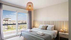 A modern hotel room with a large bed, a nightstand, a lamp, and a view of a cityscape through floor-to-ceiling windows. - New Giza’s Boho Bliss (6th of October City)