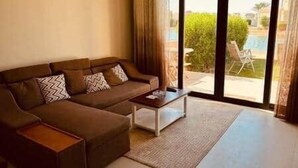 A living room with a brown sofa, a coffee table, and a view of a pool and greenery outside. - Sunset Haven in Tawila TownHouse (Hurghada)