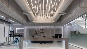 Un comptoir d’accueil moderne doté d’un habillage en verre, d’un mur métallique sur lequel les lettres « LAZ » sont clairement affichées, et d’un aménagement intérieur épuré et minimaliste.