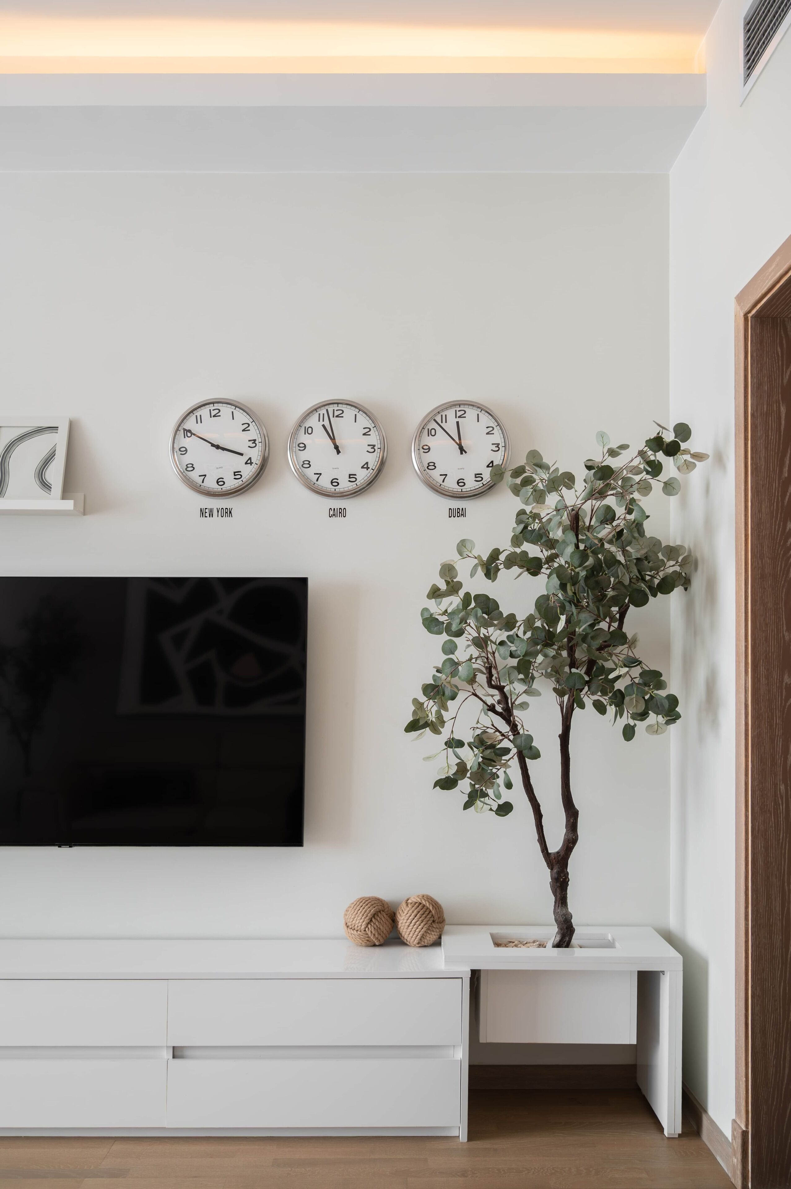 Uma sala de estar moderna com uma TV de tela plana, uma mesa de console branca e três relógios de parede com as legendas 'Nova Iorque', 'Cairo' e 'Dubai'.