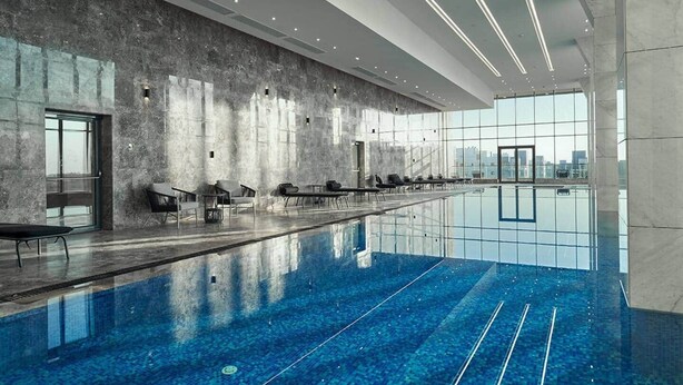 Une piscine intérieure moderne, de forme longue et étroite, avec des espaces de repos aménagés le long des parois et de grandes fenêtres offrant une vue sur la ville.